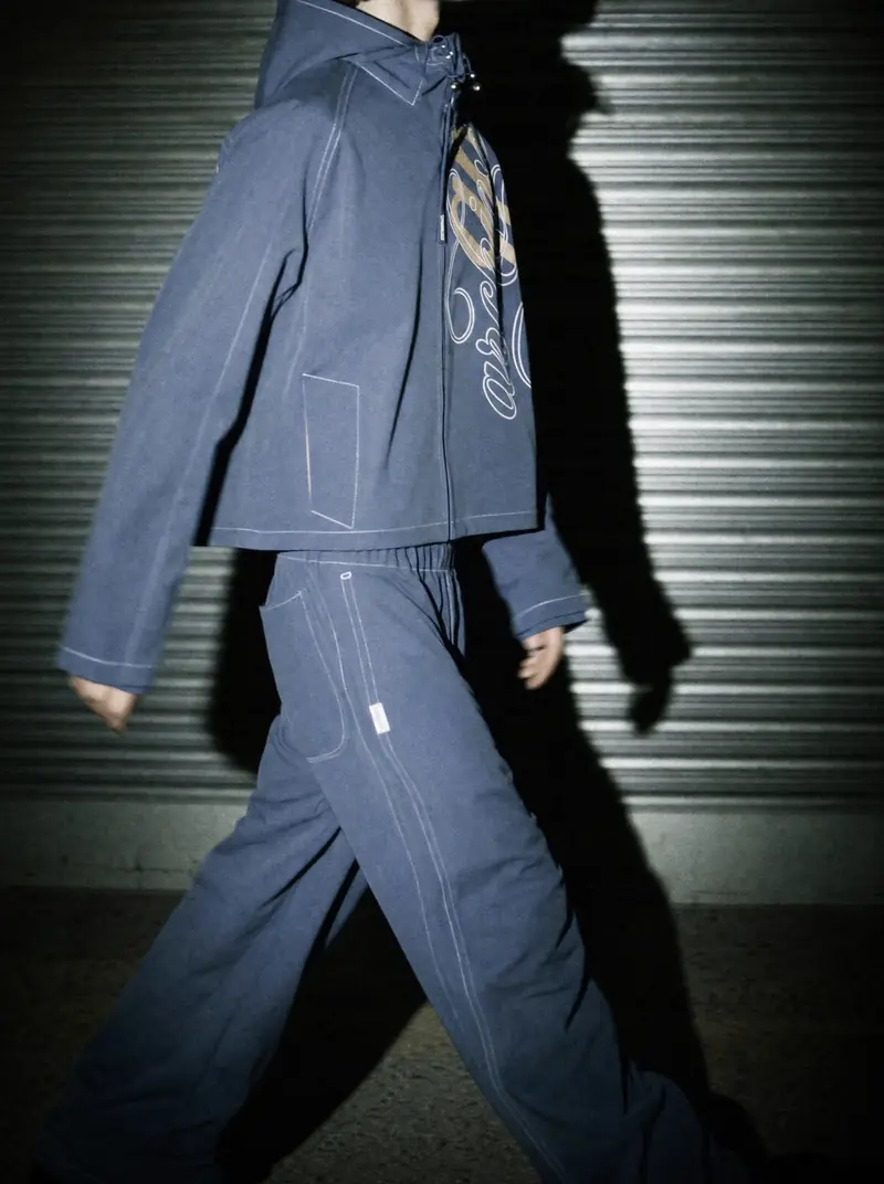 A person in a dark blue hooded tracksuit with white stitching walks past a dark corrugated roll-up door at night. A flash throws their shadow on the door. Their face is not shown.