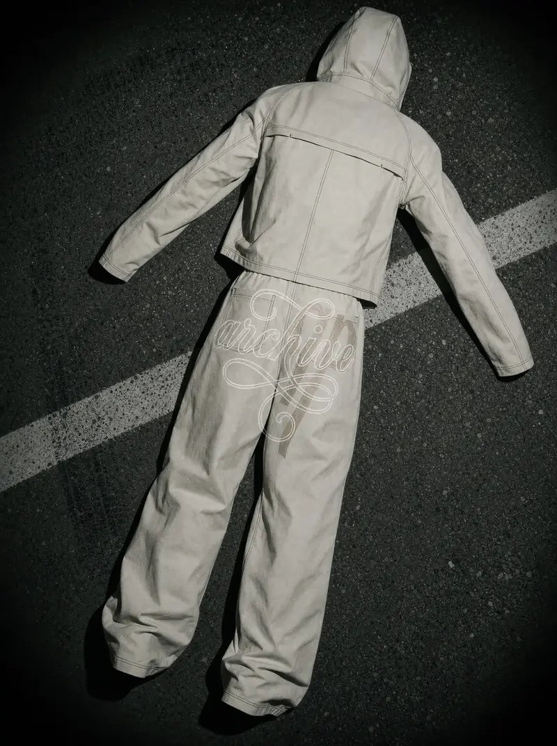 A light-coloured hooded jacket and pants laid flat on dark asphalt at night, seen from above, with a white road line behind. Large lettering and a qp mark sit across the seat of the pants.