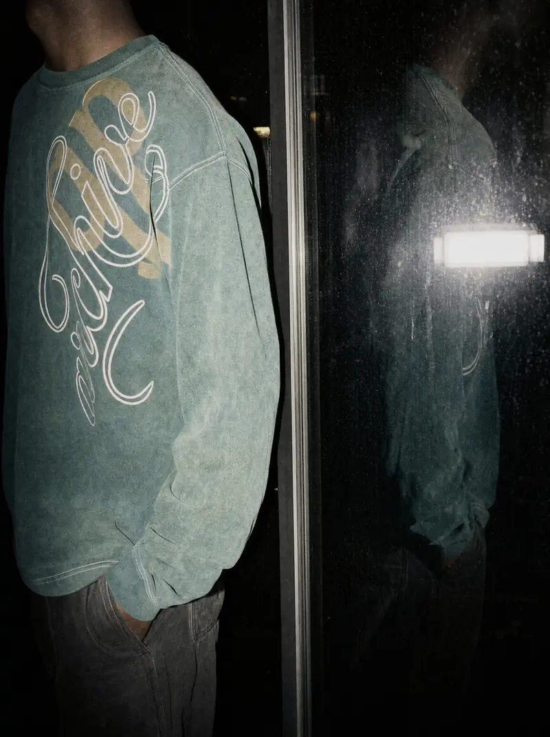 A person in a faded teal long-sleeve top stands beside a dark glass window at night, with their reflection in the glass. The top carries a white script graphic. Their face is not shown.