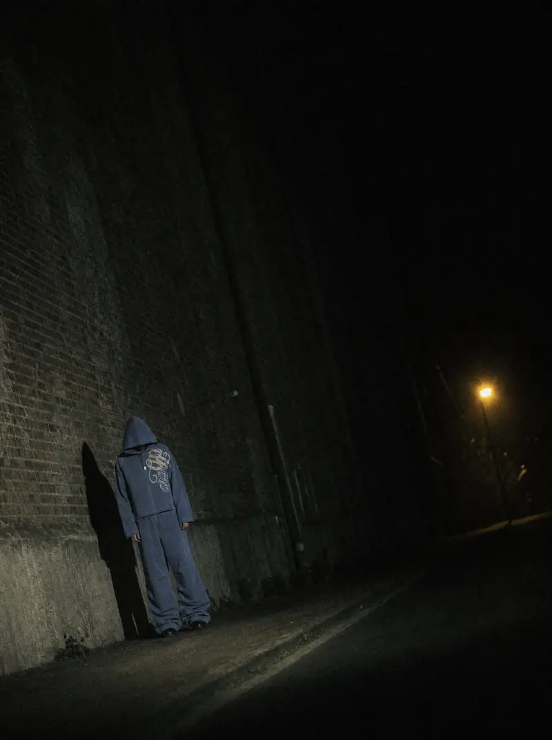 A hooded figure in a dark tracksuit stands in a dark alley at night with a streetlight glowing far behind. The hood hides their face.