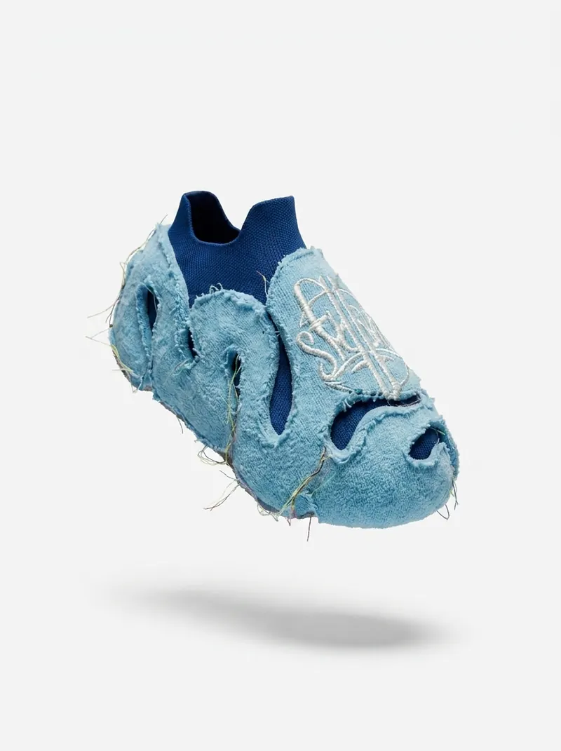 A single light blue slip-on shoe with a fuzzy outer layer, cut-outs and a white logo on top, shown at an angle on a white background.